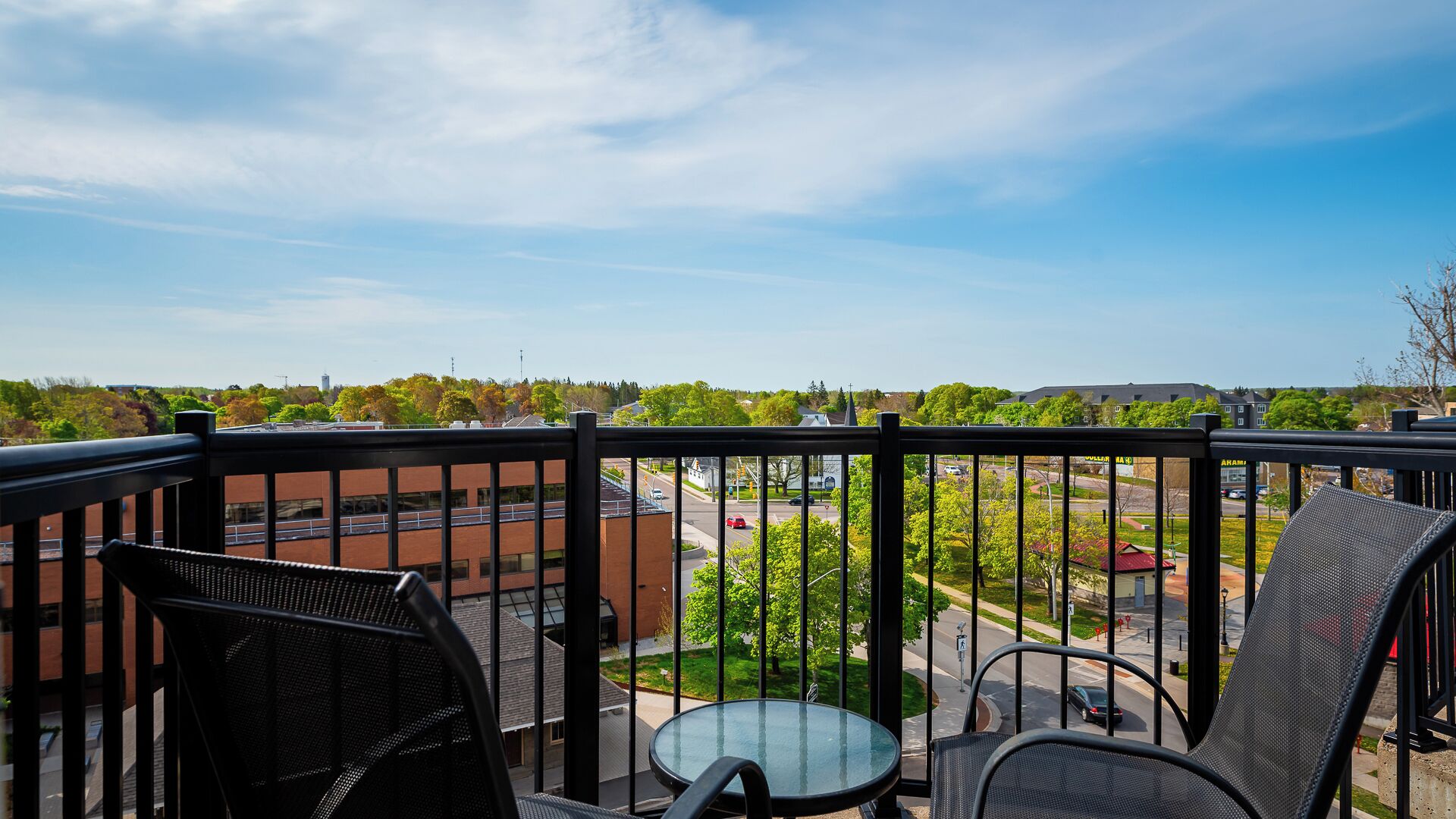 Un balcon avec deux chaises et une petite table, donnant sur une rue bordée d’immeubles et d’arbres.