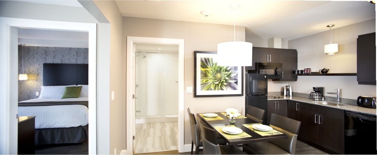 Une chambre d’hôtel moderne équipée d’un lit, d’une douche, d’un coin repas avec une table et des chaises, et d’une cuisine avec des meubles et des appareils électroménagers aux couleurs sombres.