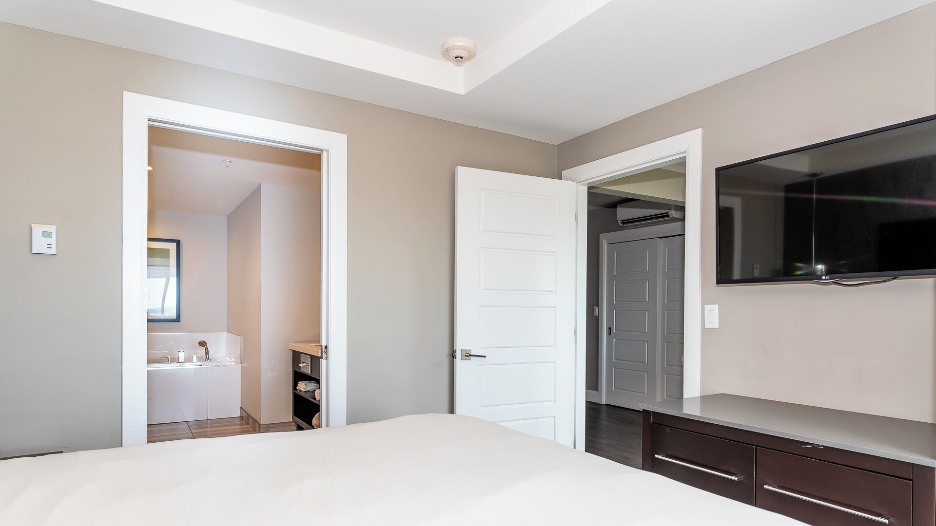 Une chambre à coucher avec un lit, un téléviseur fixé au mur, une armoire et une salle de bain visible à travers une porte ouverte.