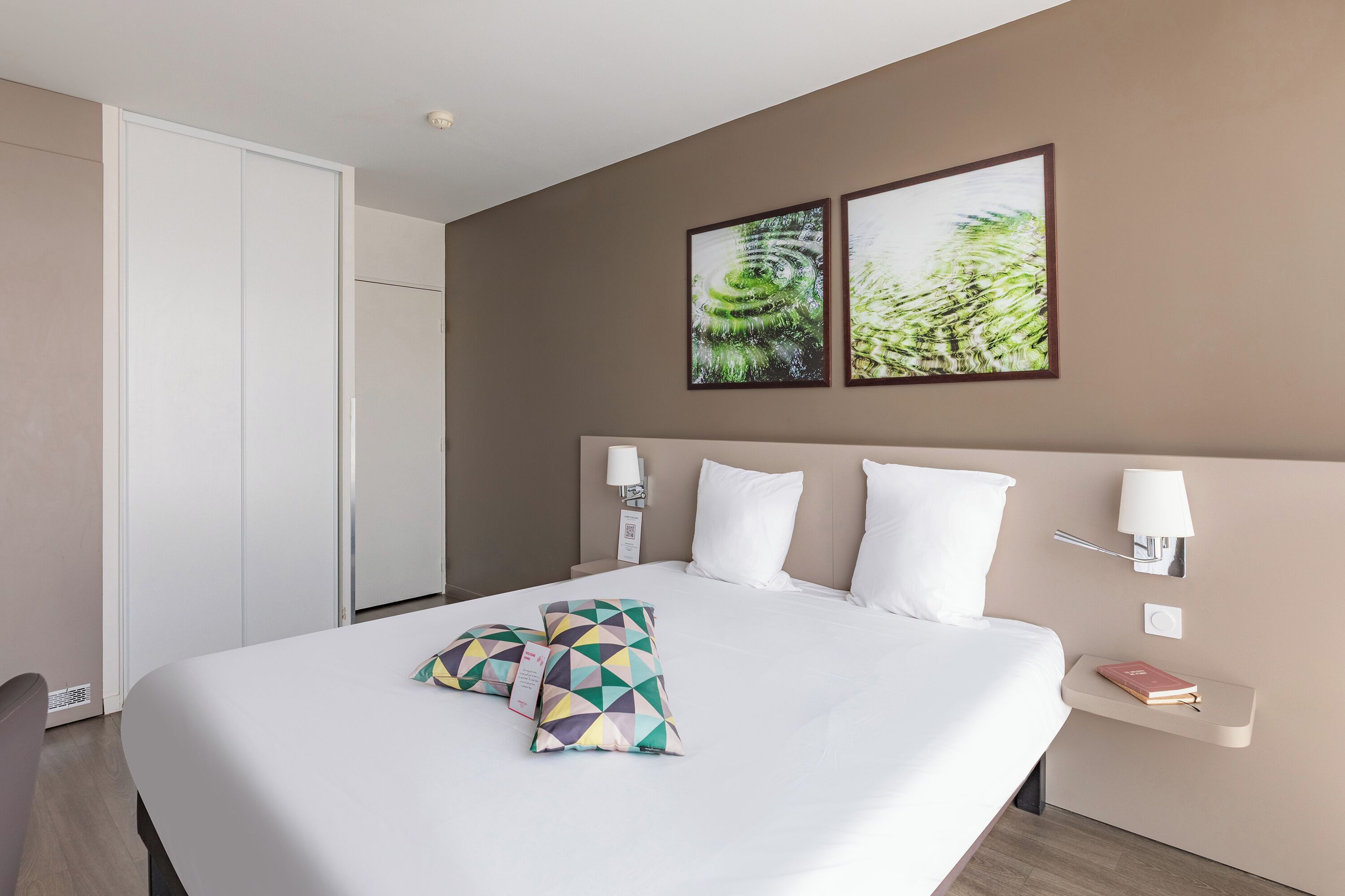 A hotel room with a large bed, two wall-mounted lamps, a bedside table with a book, and two framed pictures on the wall.