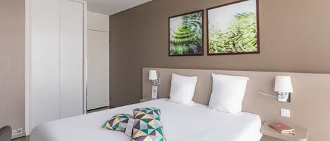 Habitación de hotel con una cama grande, dos lámparas de pared, una mesita de noche con un libro y dos cuadros en la pared.