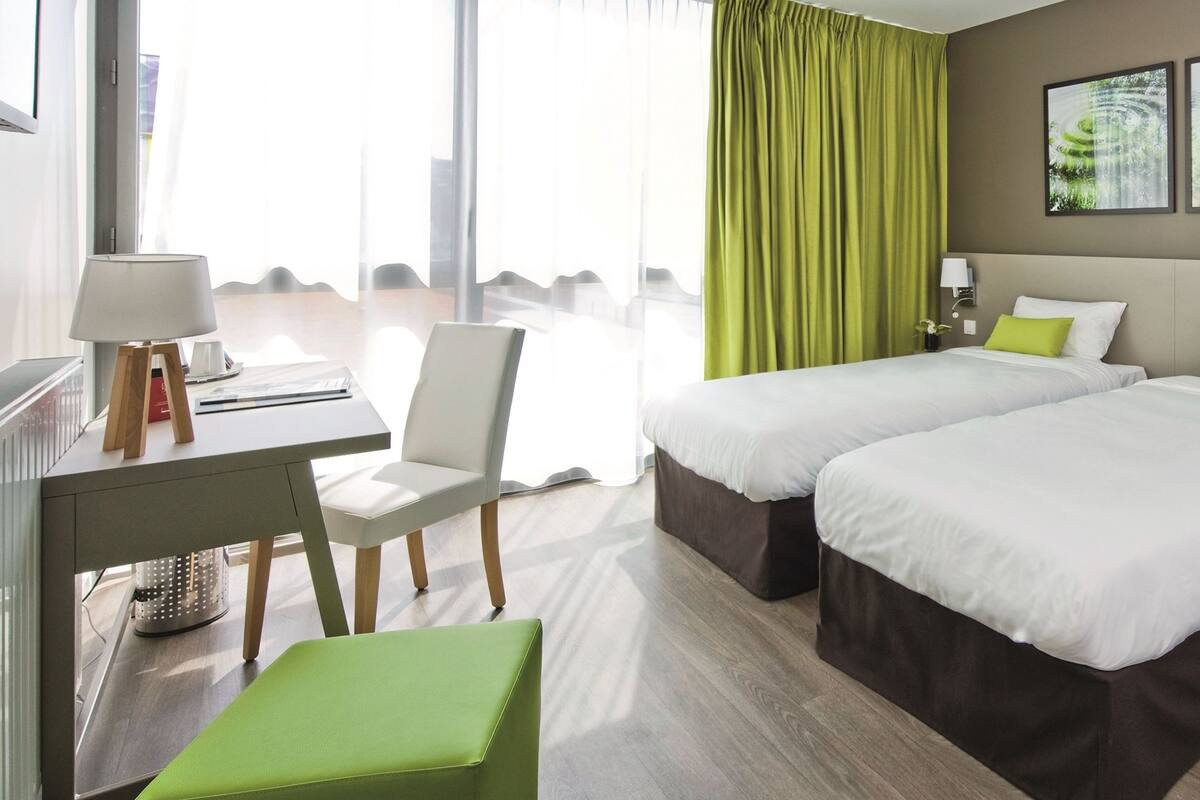 A hotel room with two beds, a desk with a chair, a small table, and a green ottoman.