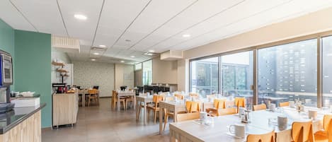 A modern dining area with multiple tables and chairs, a kitchen area with a counter and appliances, and large windows offering a view of the city.