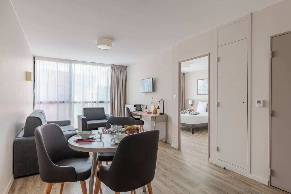 A modern hotel room with a dining area, a sofa set, a TV, and a wardrobe.