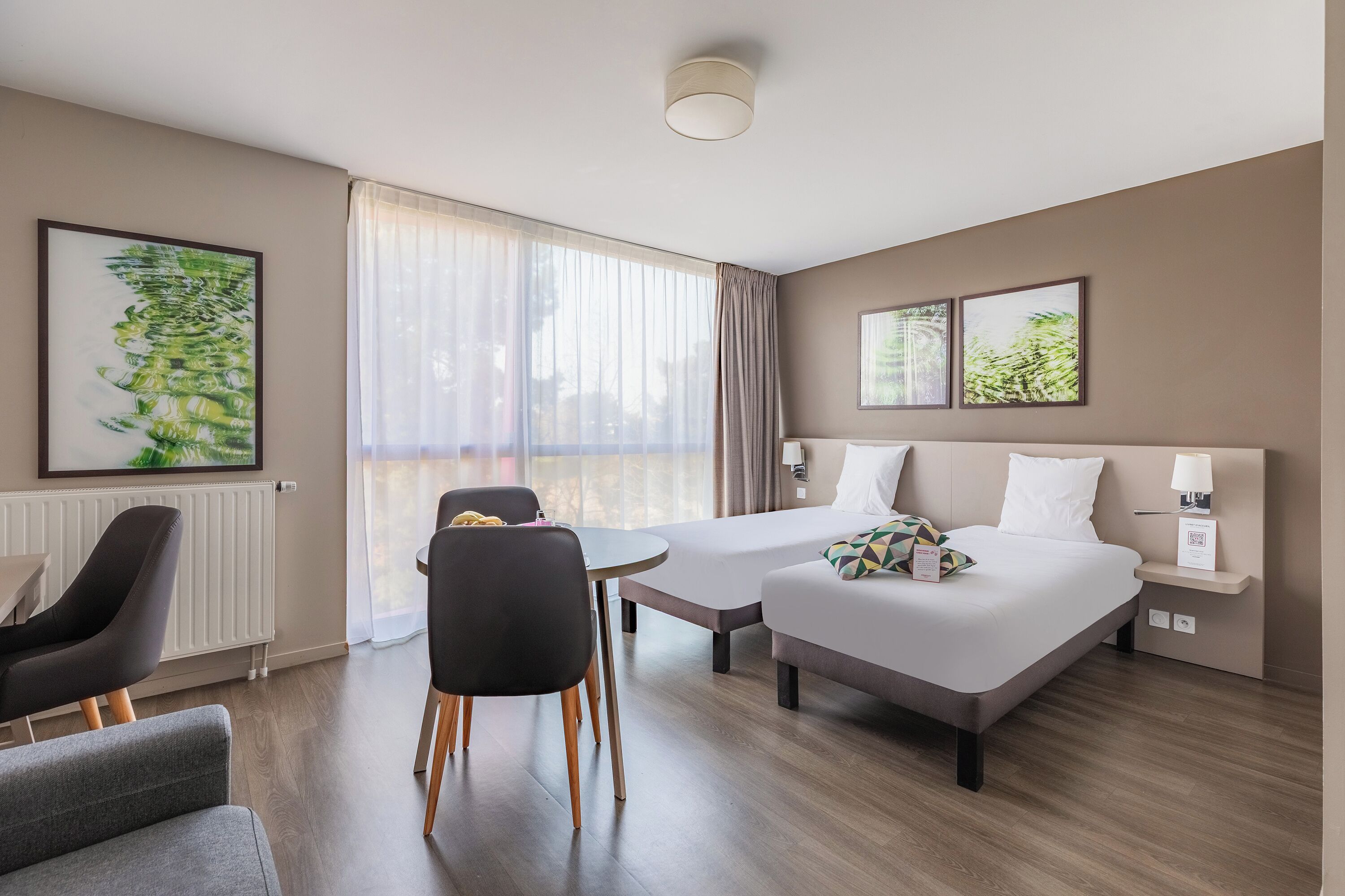 Een hotelkamer met twee bedden, een eettafel en stoelen. Er zijn grote ramen met gordijnen, een radiator en kunstwerken aan de muur.