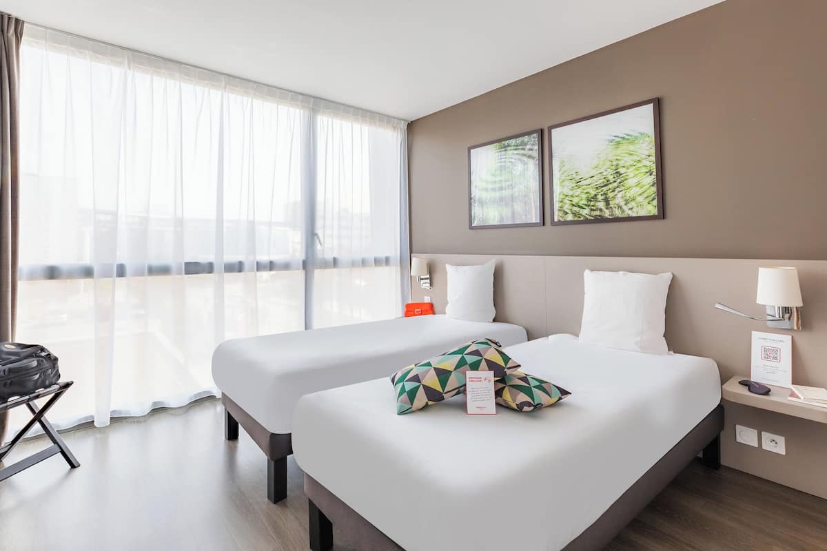 A hotel room with two beds, a large window with sheer curtains, a small table with a lamp, and a wall-mounted artwork.