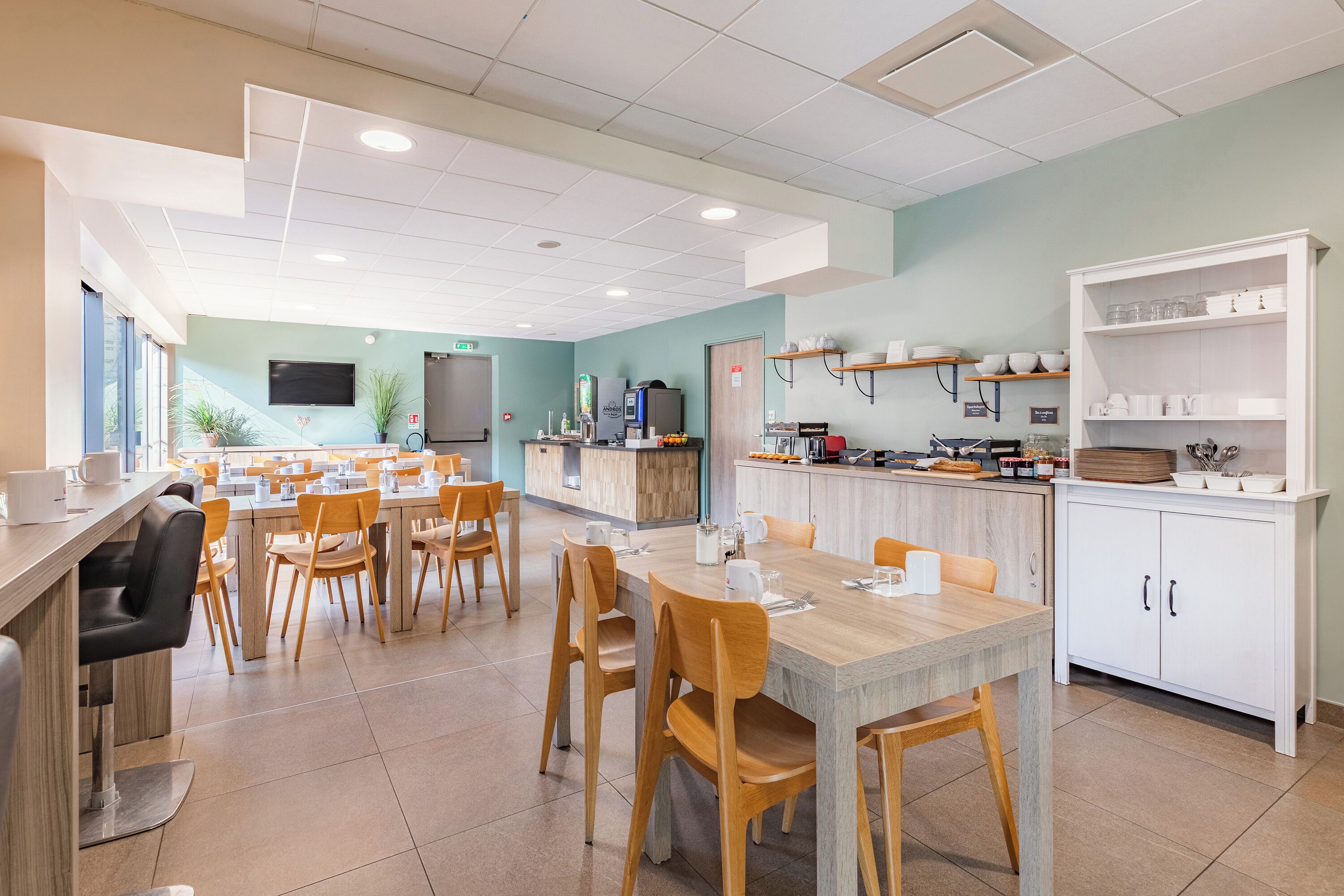 Un espace repas moderne, doté de plusieurs tables et chaises, d’un comptoir de cuisine et d’un téléviseur fixé au mur.