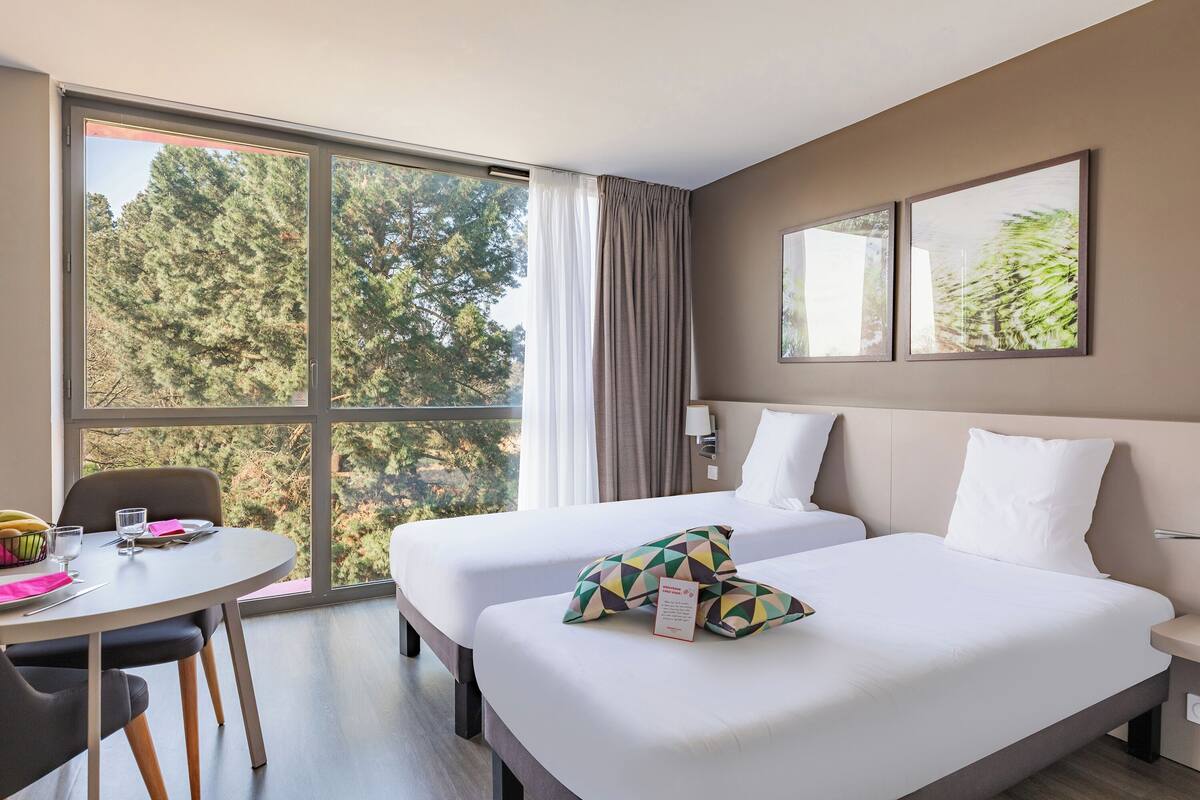 A hotel room with two beds, a dining table, and a large window with a view of trees.