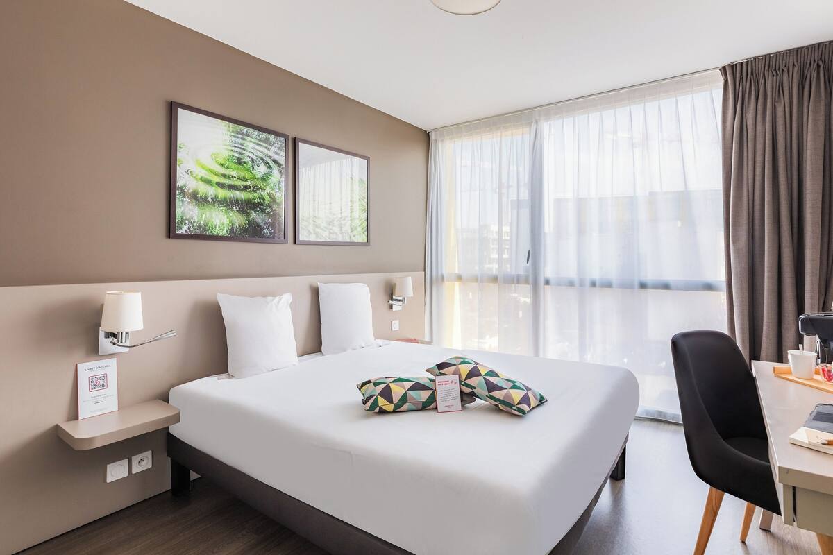 A hotel room with a large bed, a desk, a chair, and a window with curtains.