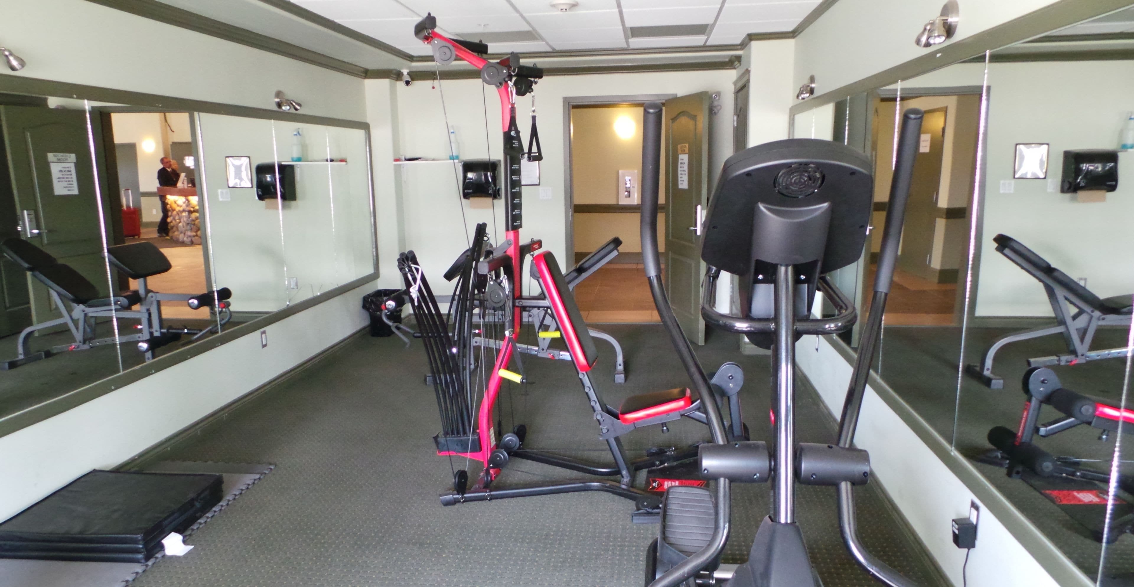 Un gimnasio con equipo de ejercicio, espejos y una puerta que da a otra habitación.
