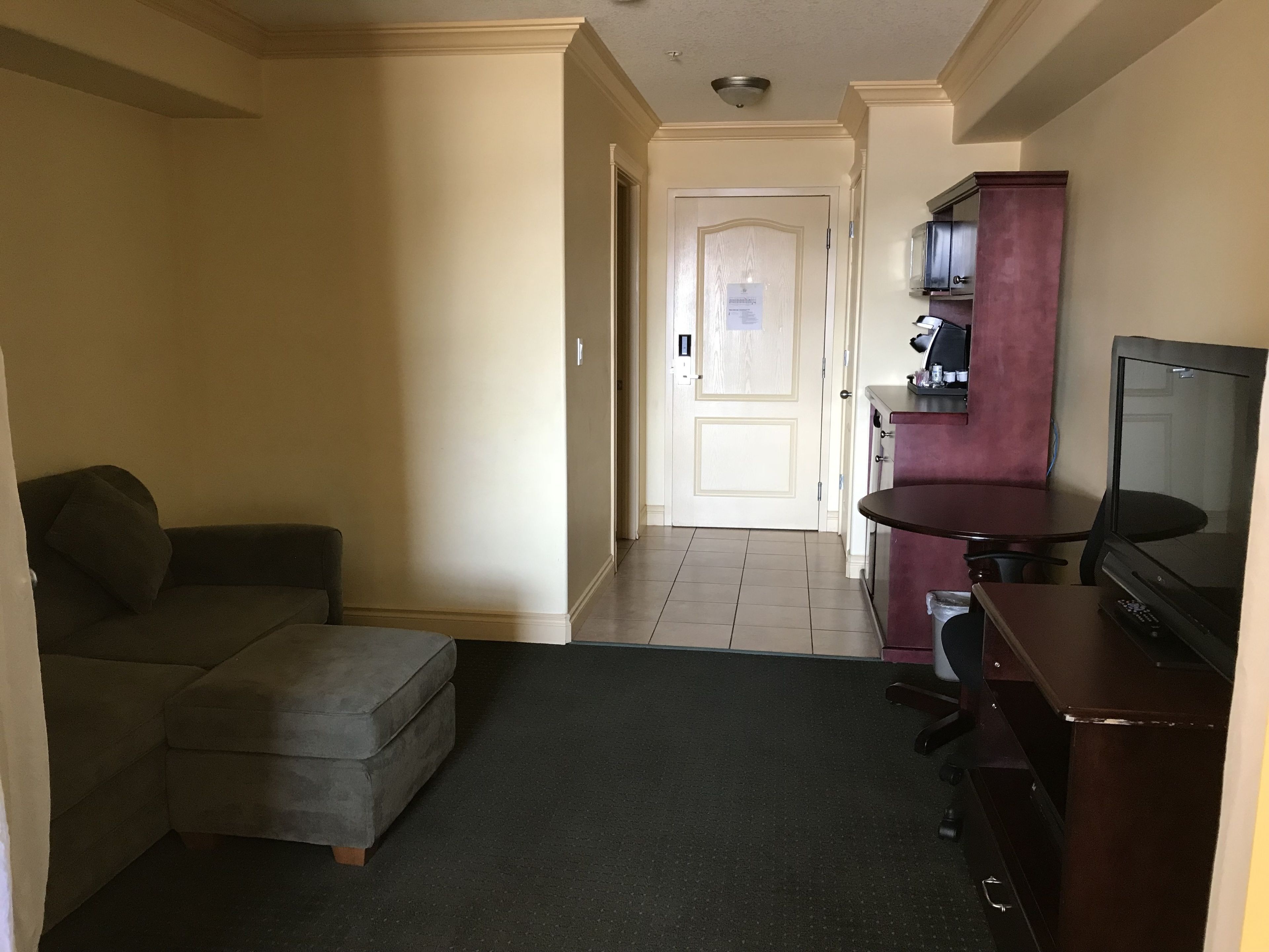 A hotel room with a sofa, a small ottoman, a desk with a chair, a television, and a minibar.