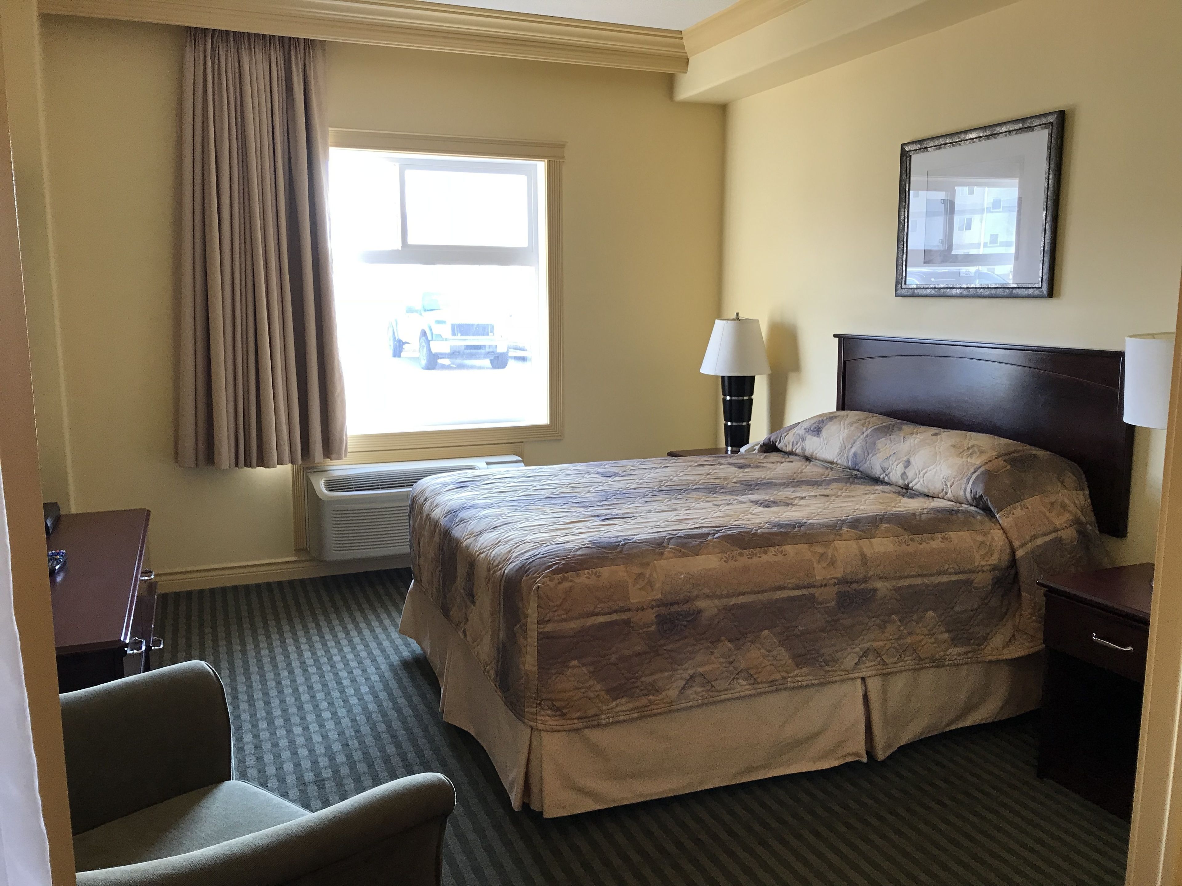 Habitación de hotel con una cama grande, un escritorio, una silla, un ventanal con cortinas, una lámpara de pared y un cuadro enmarcado.