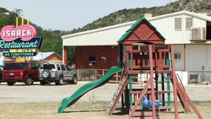 Un parque infantil con tobogán y estructura para escalar, una casita de juegos rojo y verde, y un letrero de restaurante al fondo.