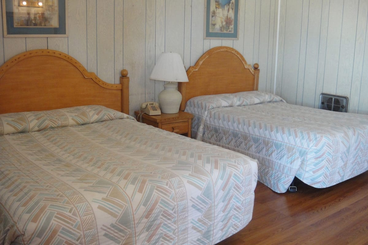 A room with two beds, wooden headboards, a bedside table with a lamp, a telephone, and a framed picture on the wall.