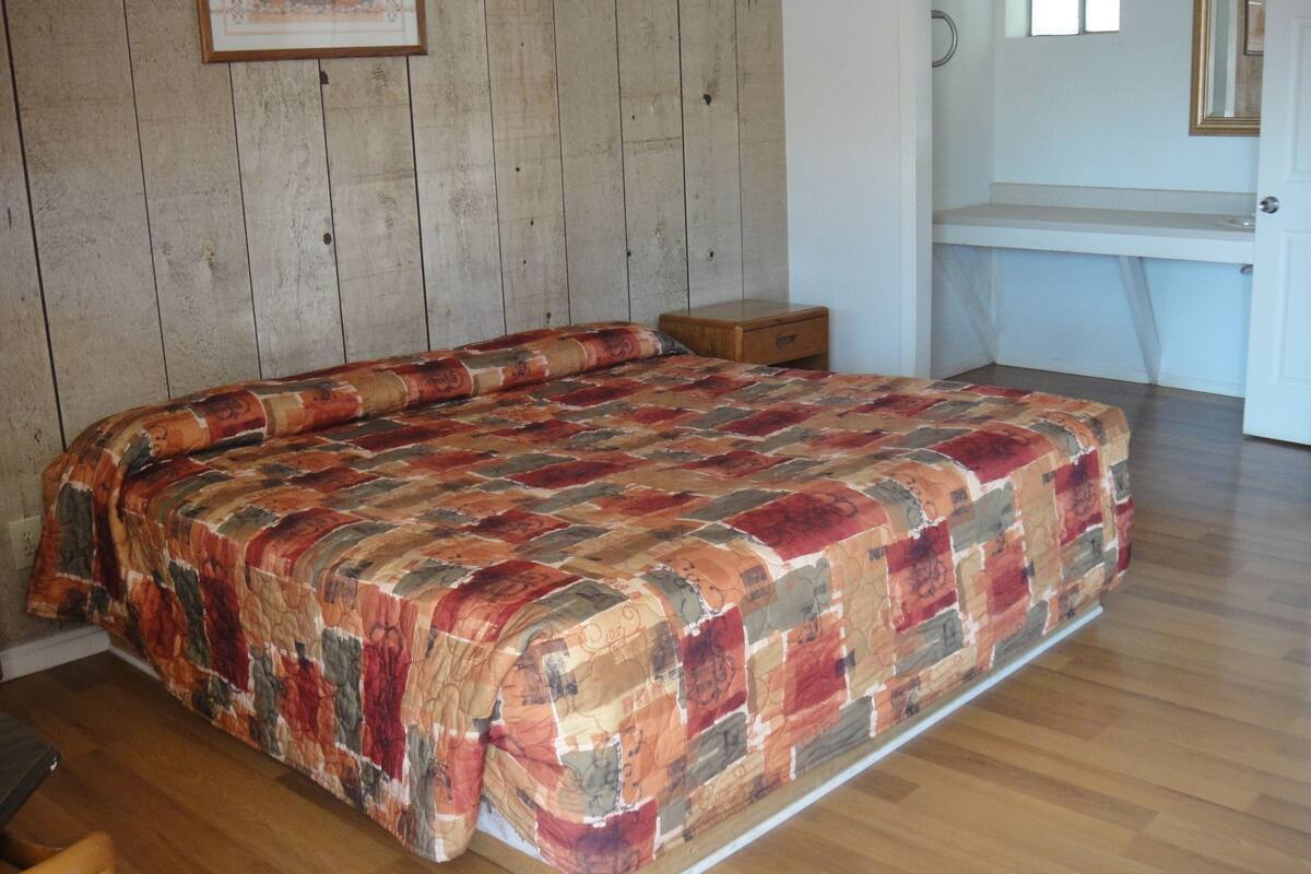 A bedroom with a wooden floor, a bed with a patterned bedspread, a nightstand, and a dresser.