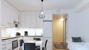 Une cuisine moderne avec des meubles blancs, un coin repas avec une table et des chaises, un réfrigérateur et un lit recouvert d’une housse de couette blanche dans une chambre.