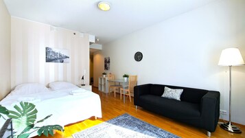 A modern, well-lit room with a bed, a black sofa, a dining table with chairs, and a small table with a lamp.