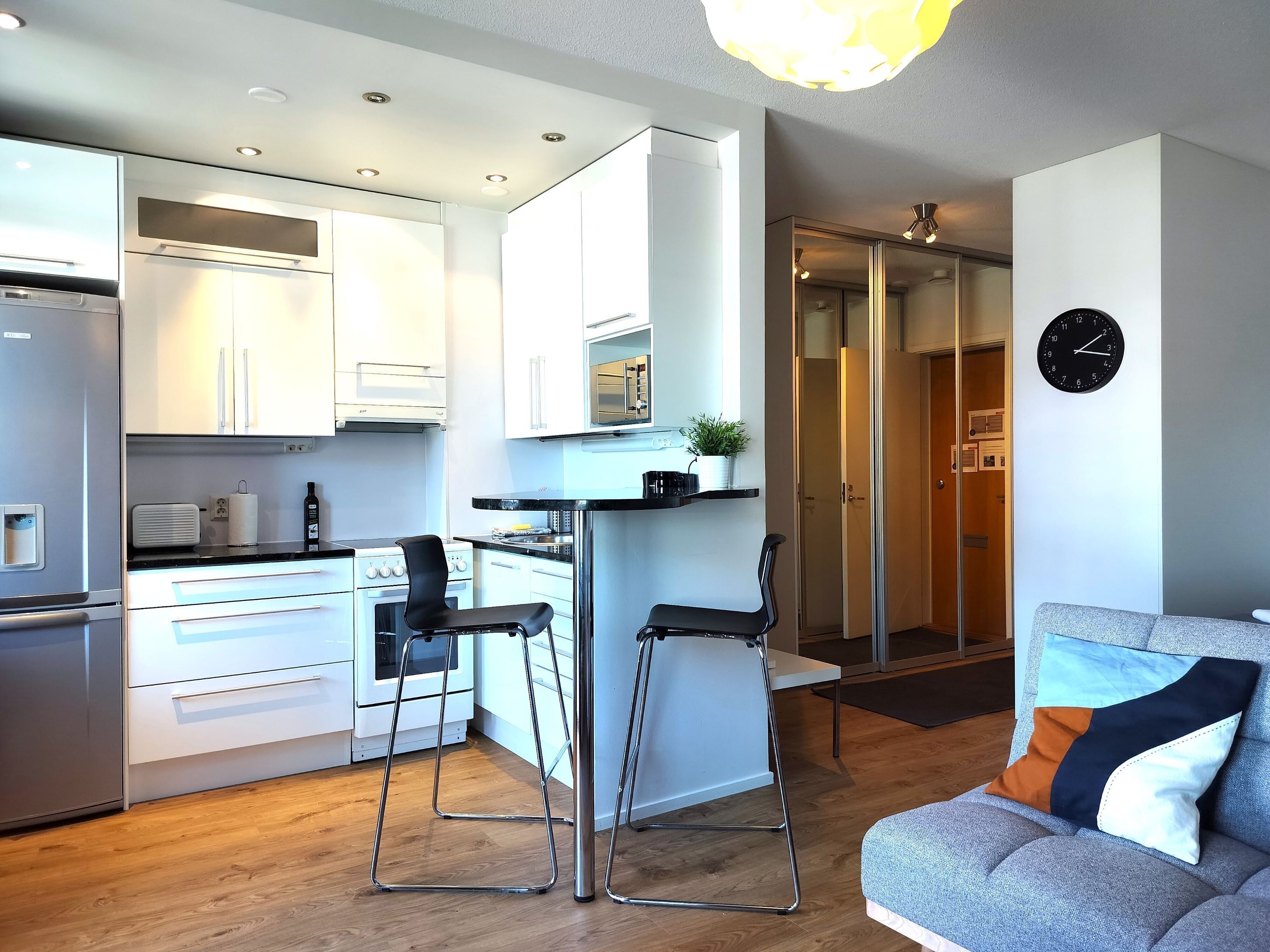 A modern kitchen with white cabinets, a stainless steel refrigerator, and a black countertop. There are two black bar stools and a gray sofa with a colorful cushion. A clock is visible on the wall.