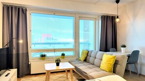 Un salon moderne avec un canapé, une petite table, un fauteuil et une grande fenêtre équipée de stores.