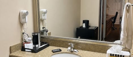Un baño de hotel con un espejo grande, un lavabo, un secador de pelo, una cafetera y un cubo de basura.