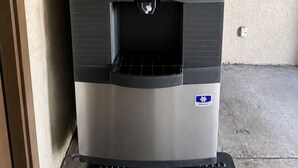 A stainless steel refrigerator with a water dispenser, located in a room with a textured wall and a concrete floor.