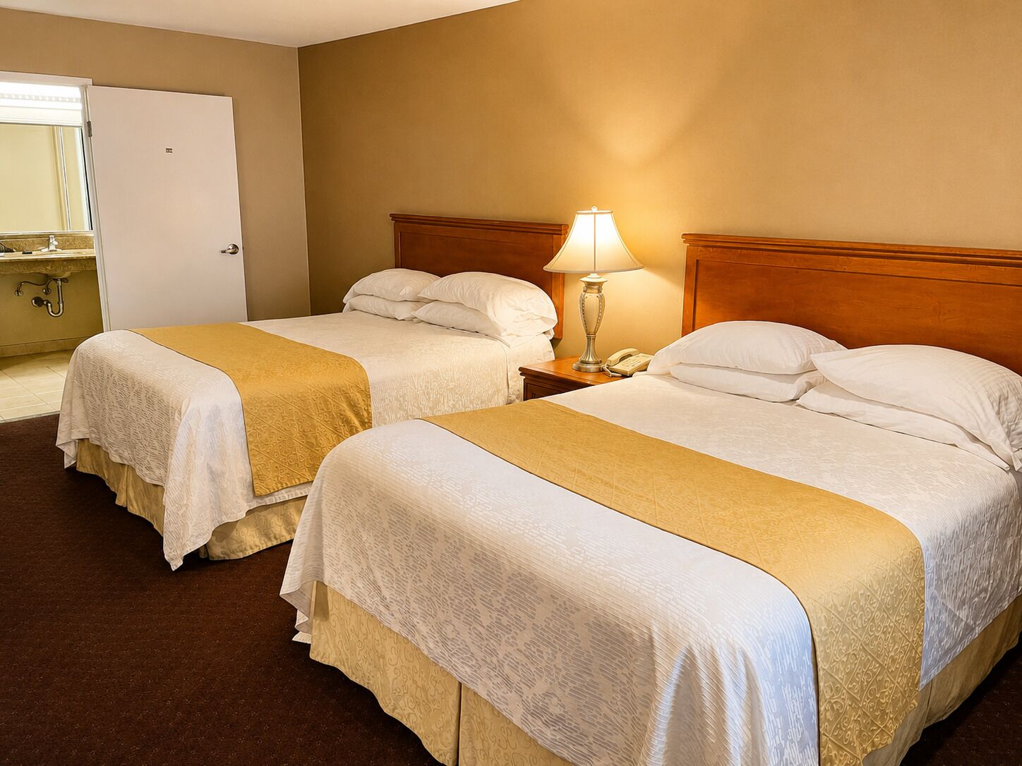 A hotel room with two beds, a bedside table with a lamp, and a bathroom with a mirror and sink.