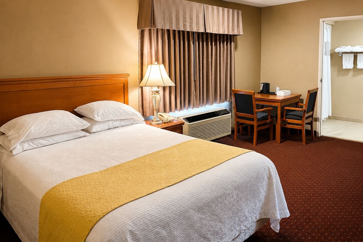 Une chambre d’hôtel avec un lit, un bureau, une chaise, une lampe et une fenêtre avec des rideaux.