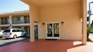 A hotel entrance with a tiled walkway leading to a double door, a parked car, and a balcony with railings.