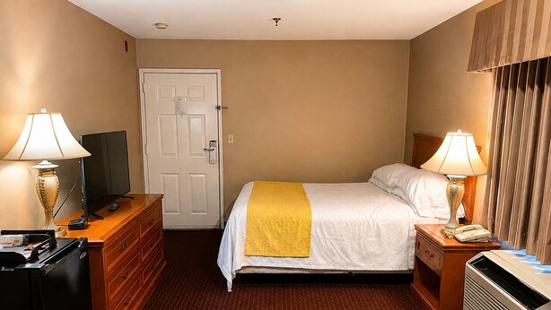 A hotel room with a bed, a dresser, a desk with a lamp, a television, and a door.