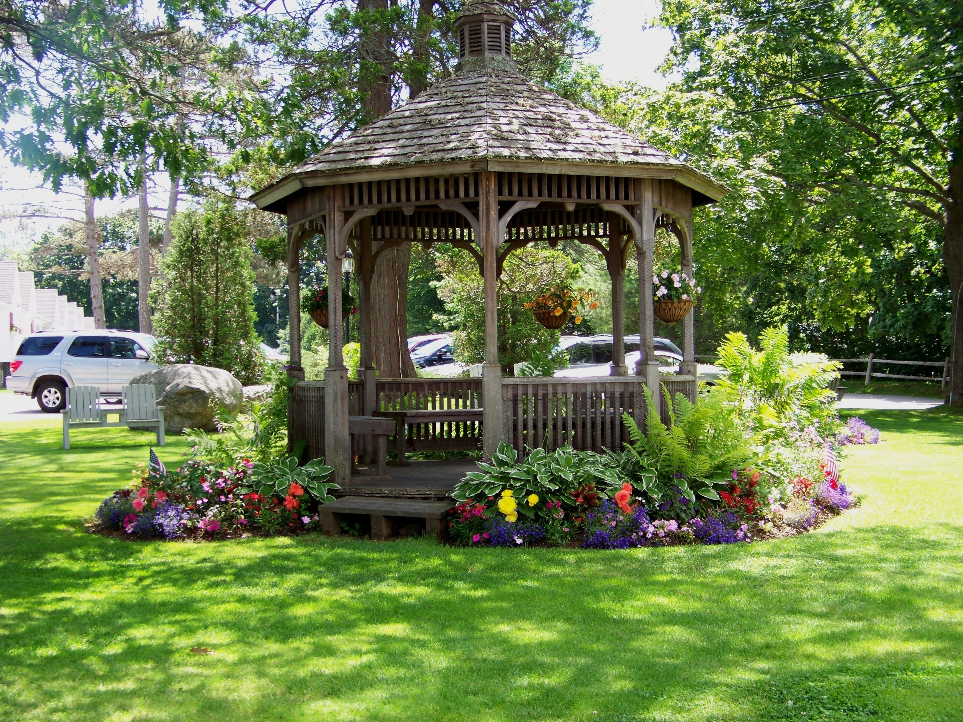 A gazebo with a wooden structure, surrounded by a well-maintained garden with various flowers and a bench.