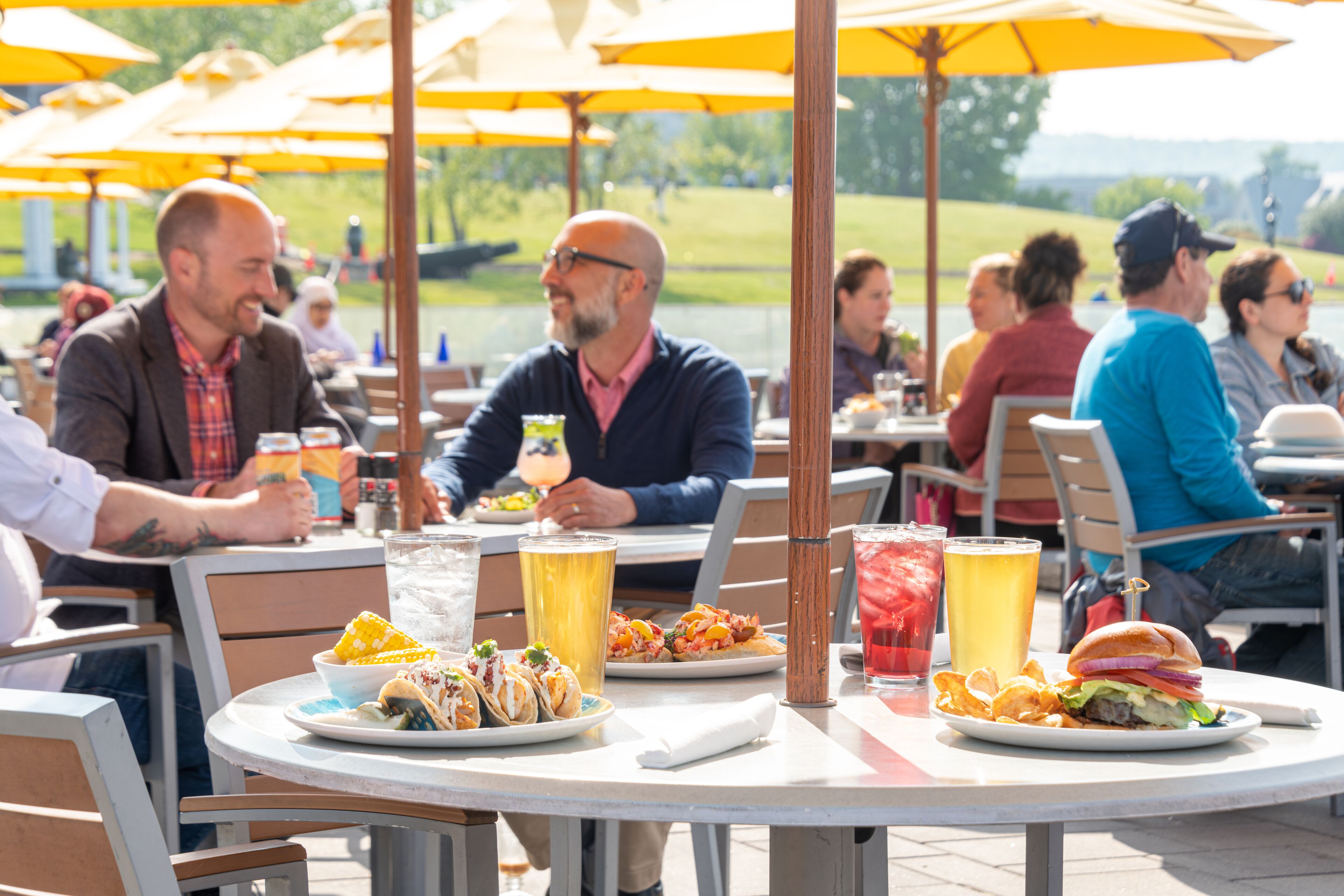 An outdoor dining area with round tables, yellow umbrellas, and various dishes including tacos, burgers, and a drink.
