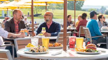 An outdoor dining area with round tables, yellow umbrellas, and various dishes including tacos, burgers, and a drink.