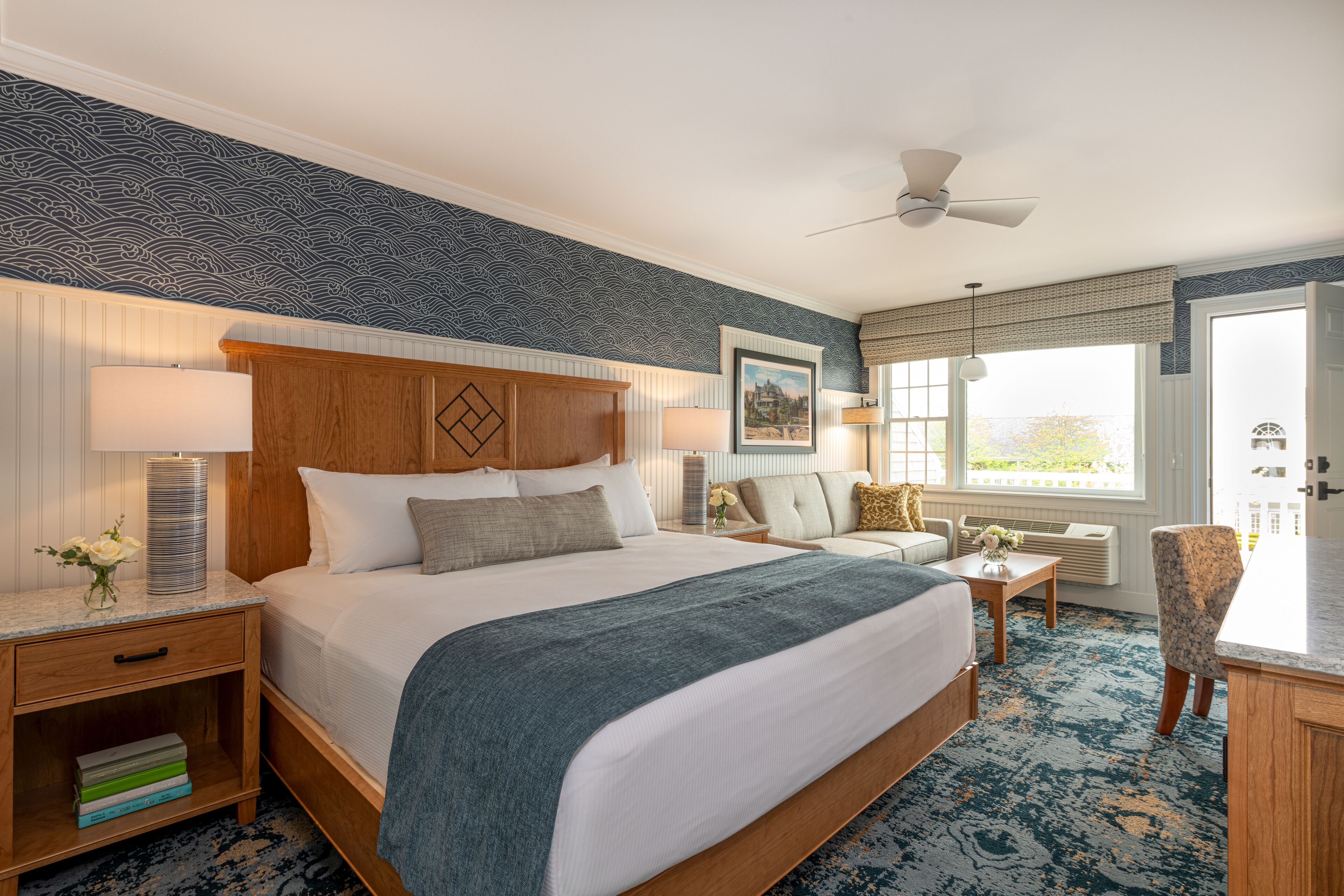 A hotel room with a large bed, a sitting area with a sofa and armchairs, a wooden nightstand with books, and a view of the outdoors through a window.
