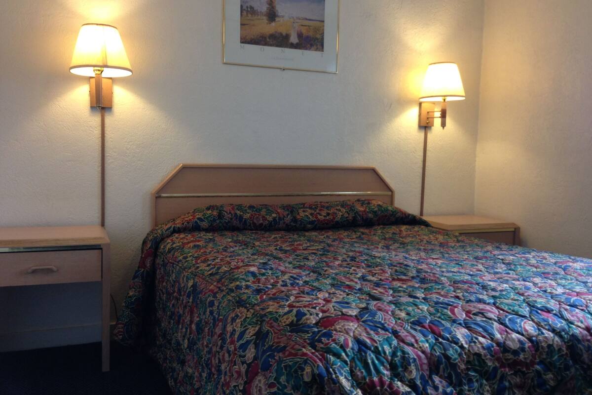 Une chambre à coucher avec une tête de lit en bois, une couvre-lit à motifs floraux, deux lampes de chevet et un tableau encadré au mur.