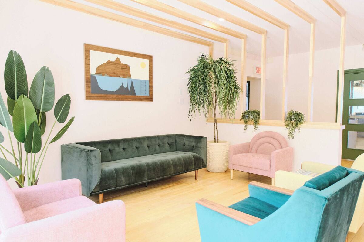 Un intérieur moderne avec un canapé vert, un fauteuil rose et des chaises bleues, une grande plante en pot et un tableau représentant un paysage, encadré.