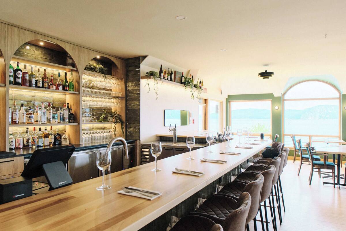 Un espace bar doté d’un long comptoir, de tabourets et d’un verre à vin. On y trouve des étagères remplies de bouteilles et de verres, ainsi qu’une fenêtre offrant une vue agréable.