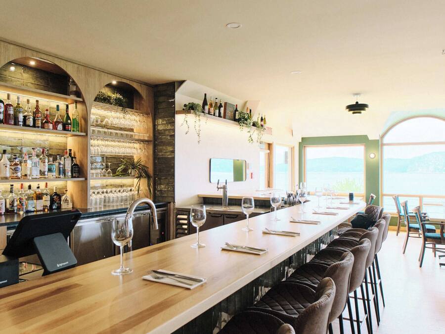 A bar area with a long counter, bar stools, and a wine glass. There are shelves with bottles and glasses, and a window with a view.