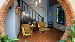 Una escalera con un arco de ladrillo que conduce a una habitación con dos sillones estampados, una mesita con un florero y un ventanal de vitral.