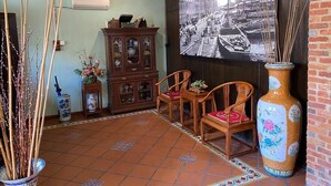 Una habitación con suelo de baldosas, un armario de madera, dos sillas de madera, un jarrón grande y un cuadro colgado en la pared.