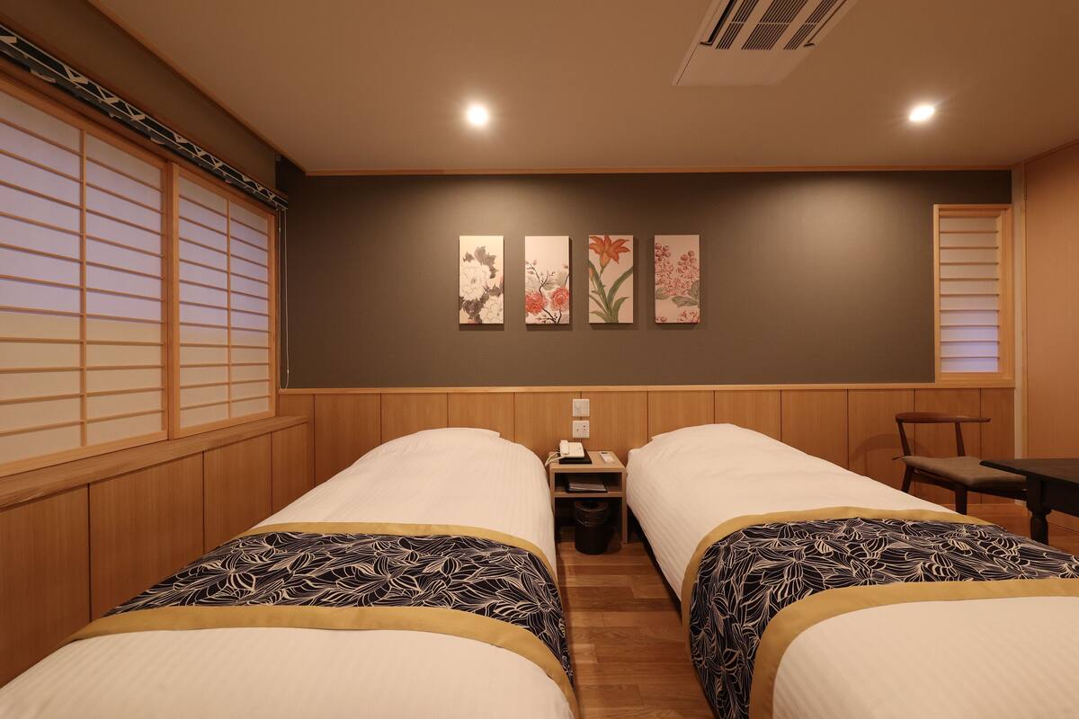 A hotel room with two beds, wooden paneling, a small table, a chair, and wall art.