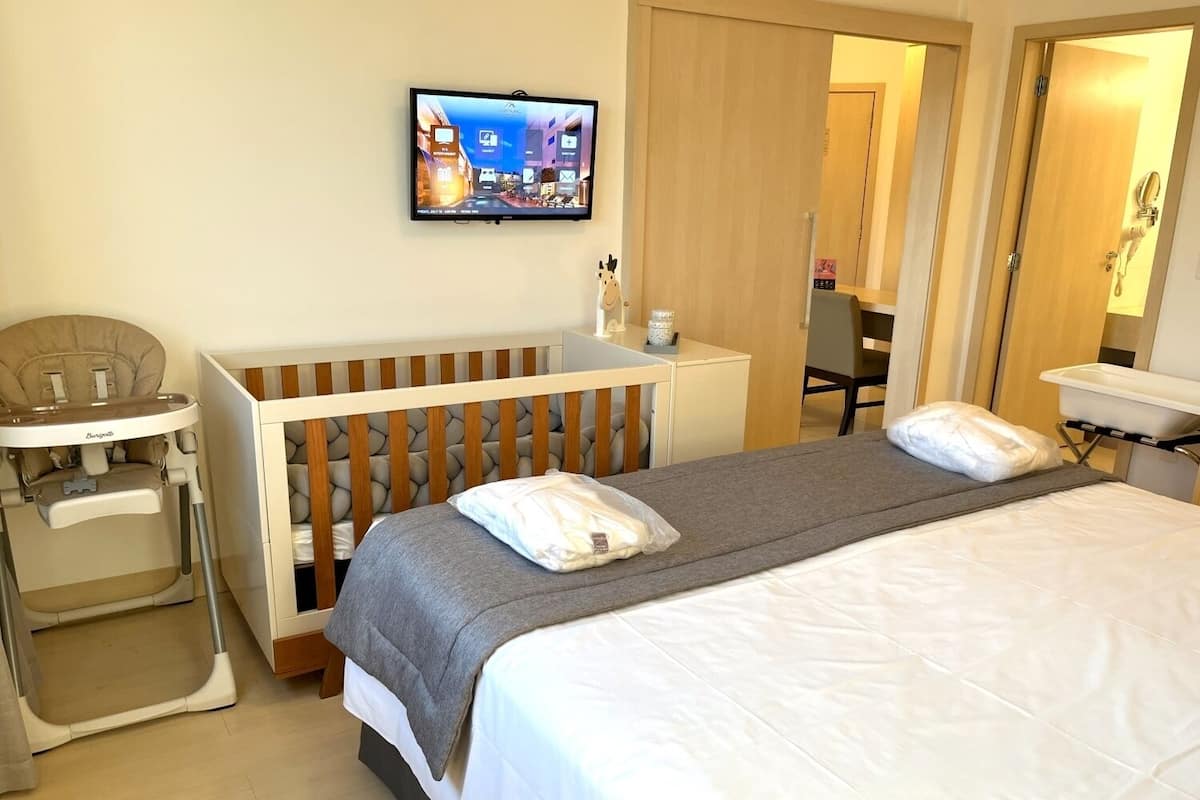 A hotel room with a bed, a high chair, a desk, and a television on the wall.