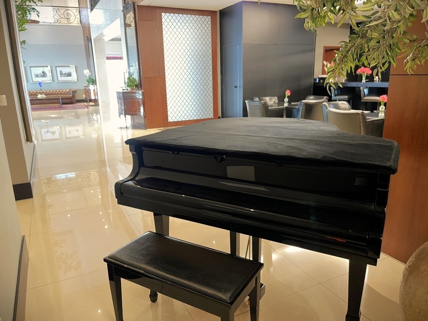 Um piano de cauda preto em um cômodo bem iluminado, com piso de azulejos, um pequeno banquinho e um vaso de planta.