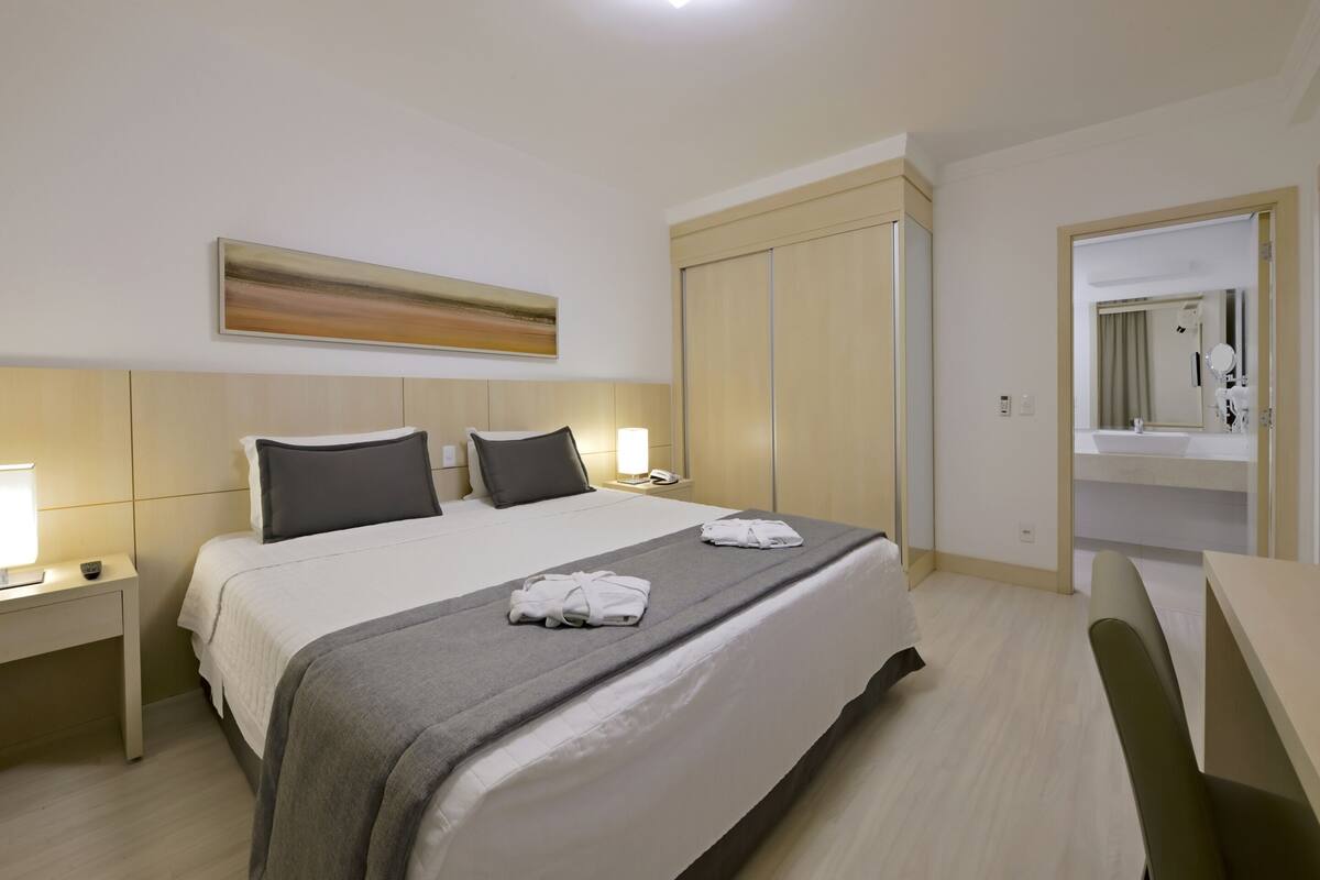 A hotel room with a bed, bedside tables, a wardrobe, a bathroom with a mirror, and a painting on the wall.