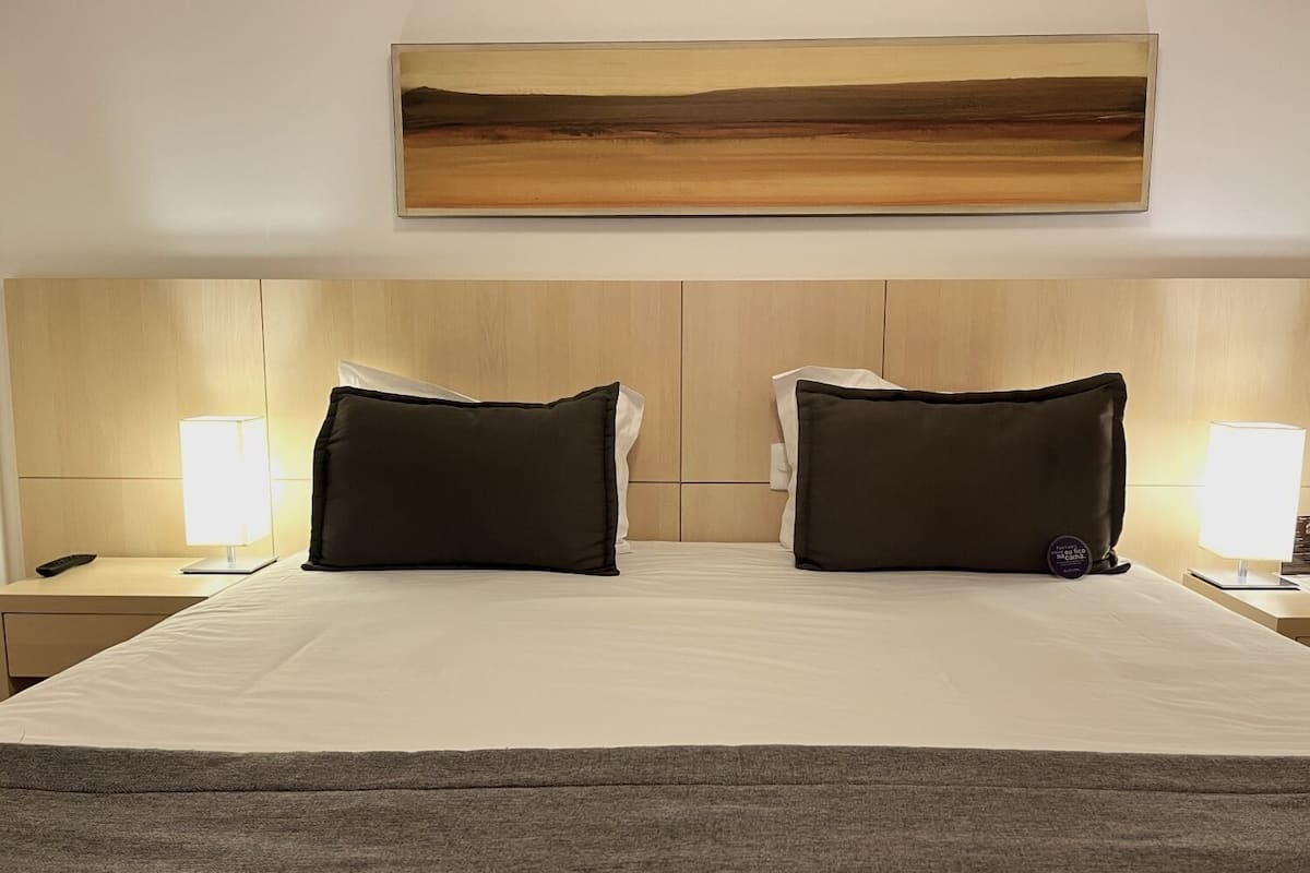 A hotel room with a large bed, two black pillows, a wooden headboard, a painting above the bed, and bedside lamps.