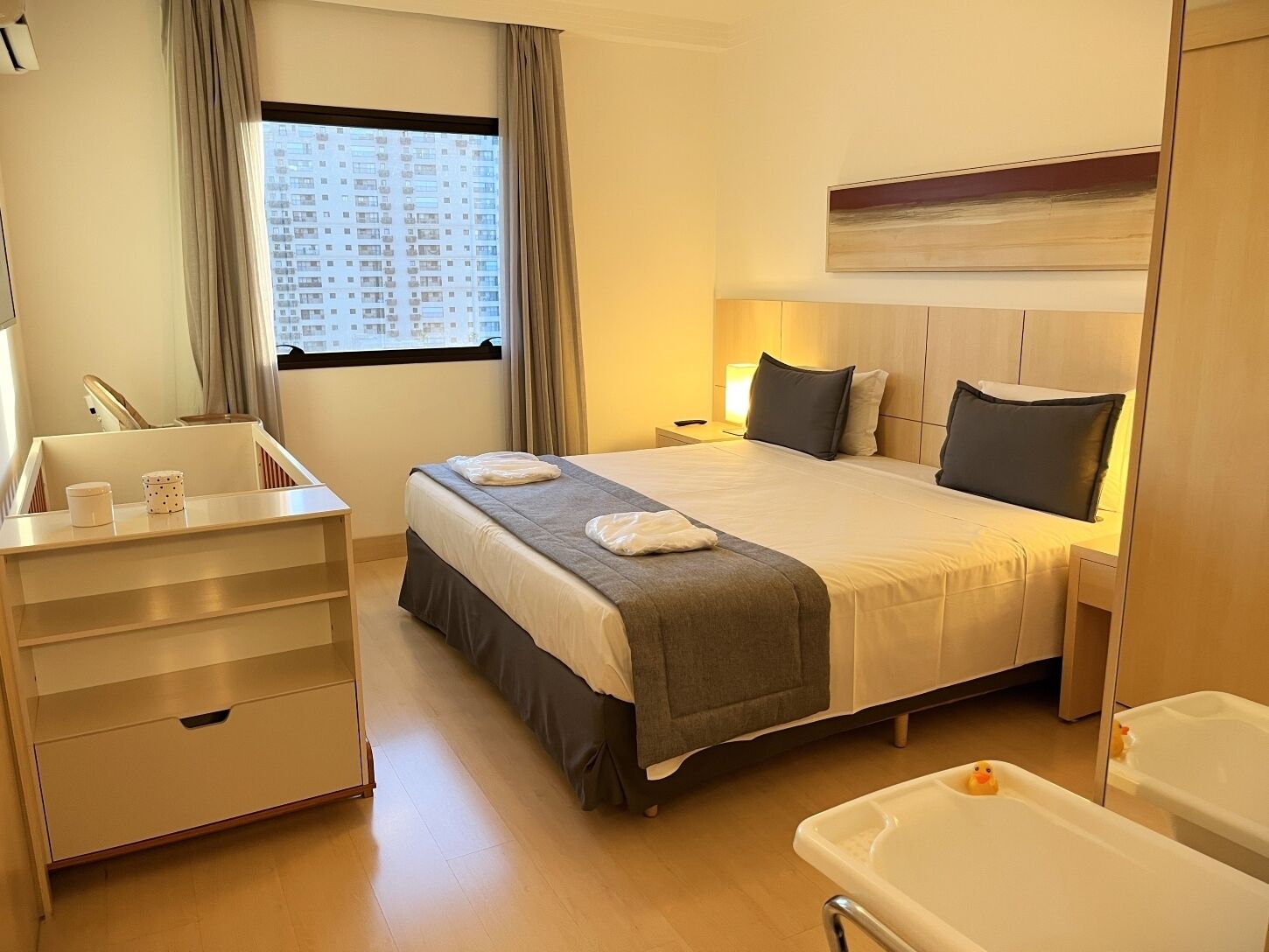 A hotel room with a bed, bedside table, lamp, and a view of a building through the window.