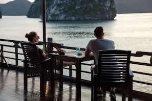 Two people seated at a table with a view of a body of water and a distant island. - Halong Glory Cruise (Ha Long)