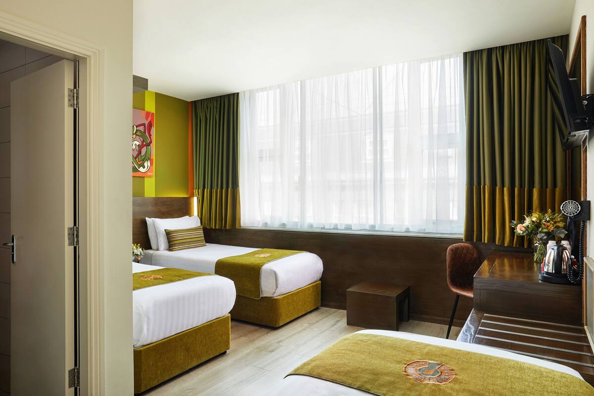A hotel room with two beds, a desk, a chair, and a large window with sheer curtains.