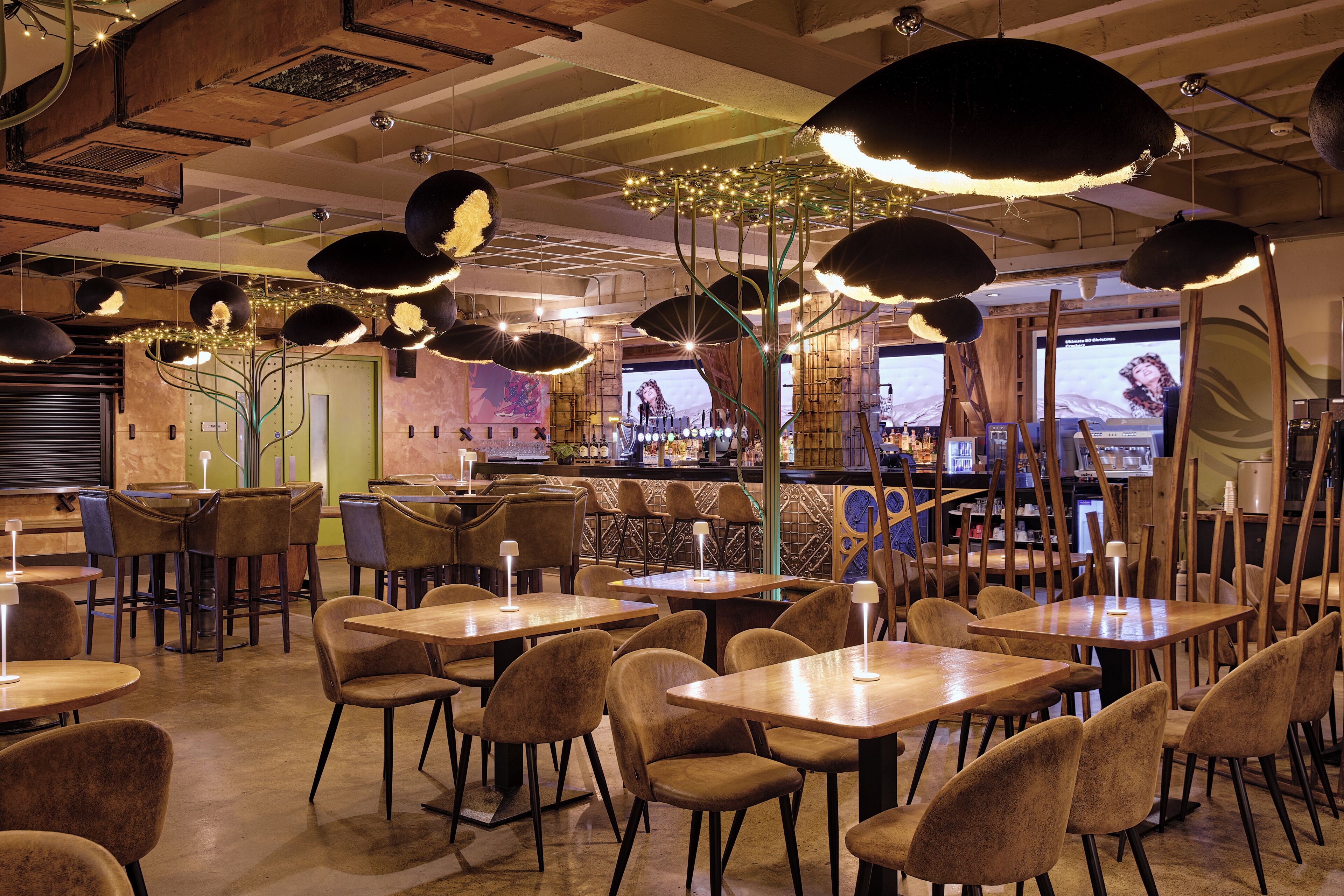 A restaurant interior with multiple tables and chairs, pendant lighting, and a bar area.