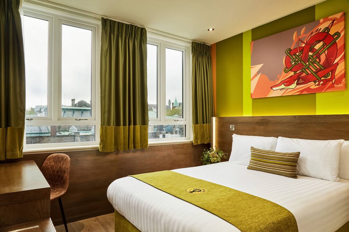 A hotel room with a bed, a desk, and a view of a cityscape through large windows.
