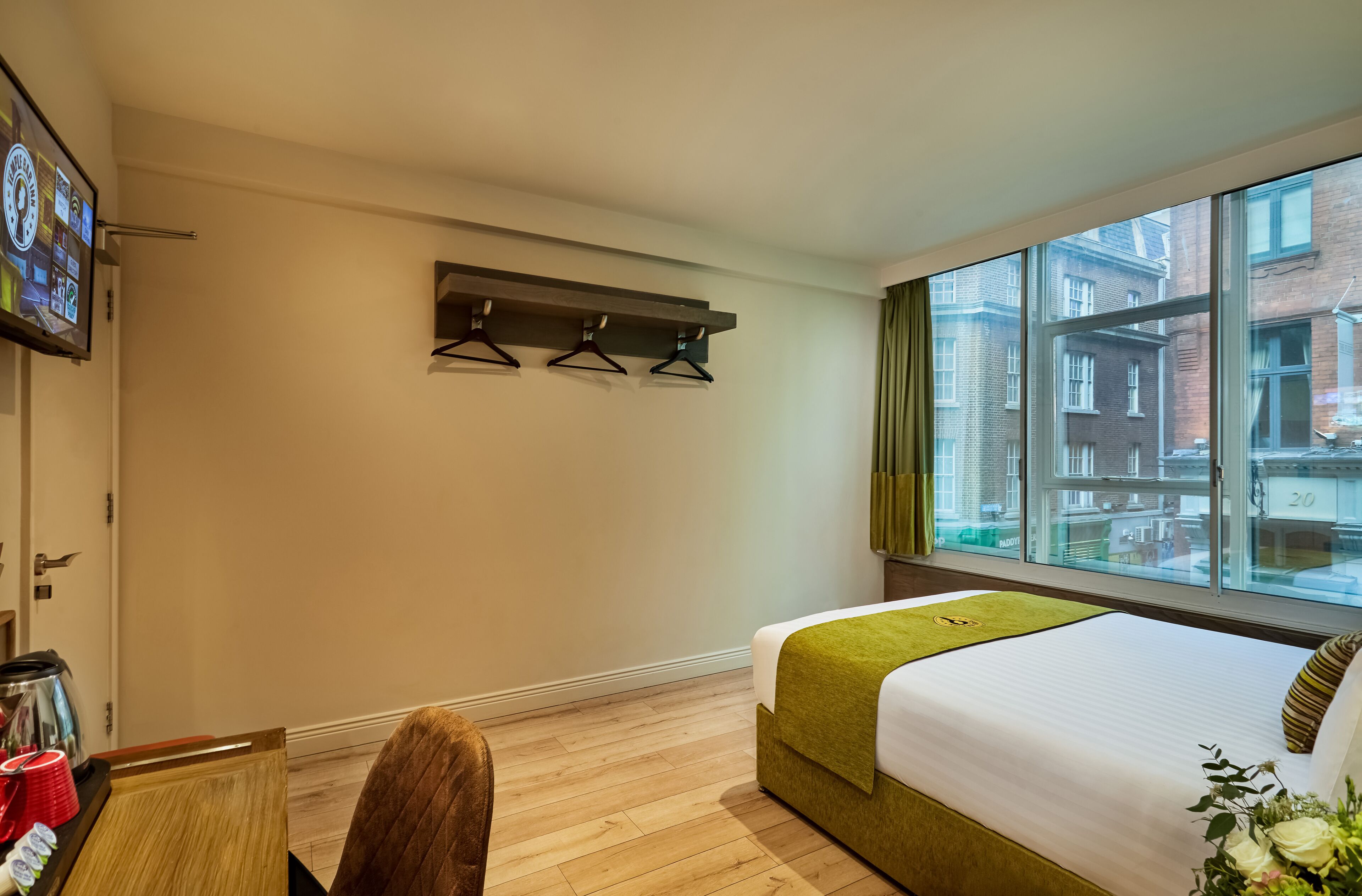 A hotel room with a bed, a TV mounted on the wall, a window with a city view, and a small table with a kettle and cups.